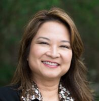 A woman with long brown hair, wearing a black blazer and patterned blouse, stands outdoors with her arms crossed, smiling at the camera.