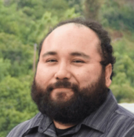 A man with a beard and mustache, wearing a dark striped shirt, stands outdoors in front of green foliage.
