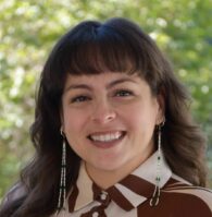 A woman with long brown hair and beaded earrings smiles outdoors, wearing a brown and white patterned blouse. Green foliage is visible in the background.