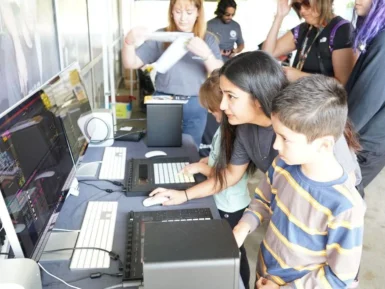 People engaged in working on computers, including a woman and a child, surrounded by others at a tech setup with large monitors.