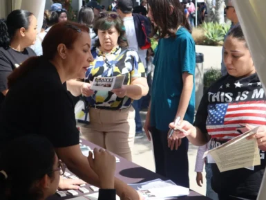 People engaged in a discussion at a booth, one holding pamphlets, others interacting. Some are standing, while one is seated. The booth has flyers and giveaways spread on it.