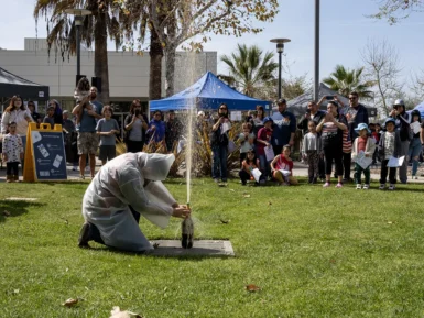 A person in protective gear kneels on grass, creating an erupting fountain with a soda bottle. A crowd watches, standing by blue tents and palm trees in the background.