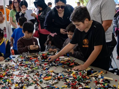 Children and adults gather around a table filled with various Lego pieces, with some actively building and others observing.