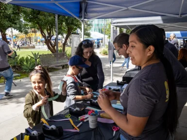 Children and adults engage in arts and crafts at a booth under a canopy. Tables are filled with colorful paper and supplies. Other people and booths are visible in the background.