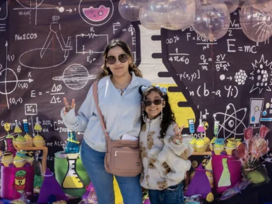 Two people pose in front of a science-themed backdrop with colorful decorations and desserts.