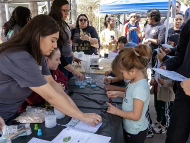 People gather around a table for a science activity, with children participating in experiments and adults assisting under a tent.