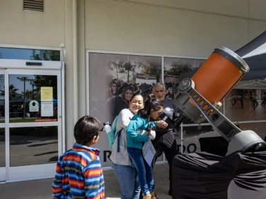 A woman helps a child look through a large telescope outside a building while another child watches.