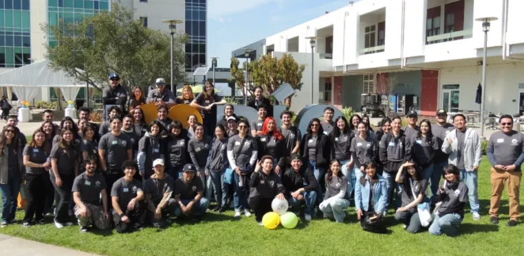 A large group of people poses for a photo on grass in front of modern buildings during the day; some hold balloons and signs.