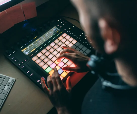 Person using a grid-based music production controller next to a keyboard and a computer screen.